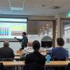 View of a speaker talking to slides at the front of the room.